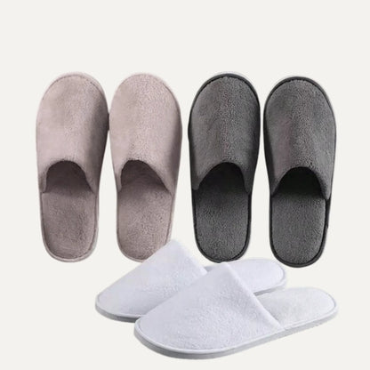 Three pairs of plush slip-on slippers shown on a plain light background: two in muted taupe and one in dark charcoal, with a third pair in white placed in front.