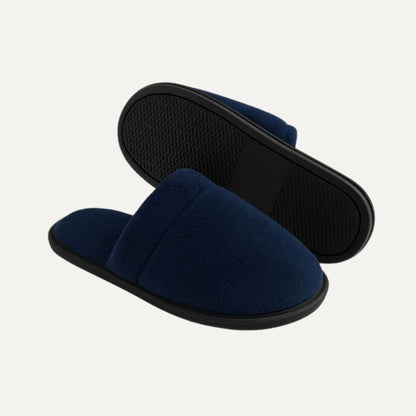 A pair of navy blue slip-on slippers with black soles, shown on a light off-white background. One slipper is positioned in front of the other to show the textured black outsole.