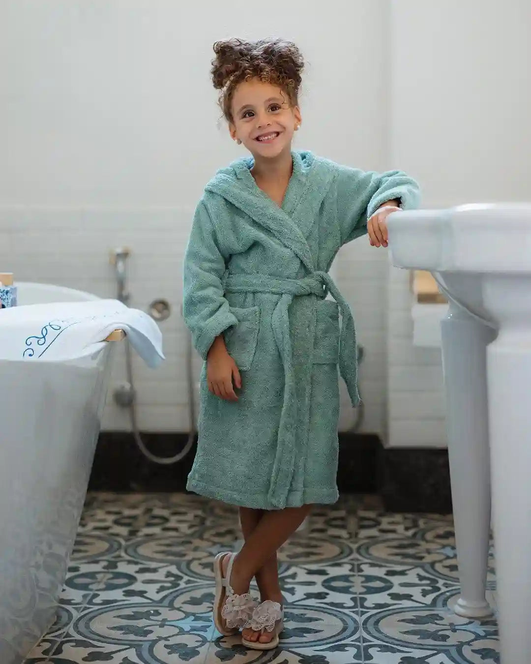 A young girl with curly brown hair stands in a bathroom wearing a plush mint green bathrobe and white slip-on shoes, leaning against a white pedestal sink.
