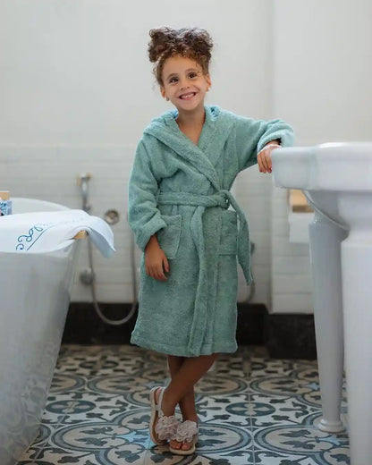 A young girl with curly brown hair stands in a bathroom wearing a plush mint green bathrobe and white slip-on shoes, leaning against a white pedestal sink.