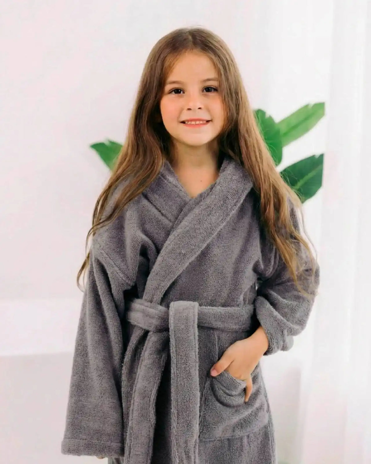 A young girl with long brown hair smiling while wearing a plush gray bathrobe with a tie belt, standing against a white curtain background with a green plant leaf visible behind her.